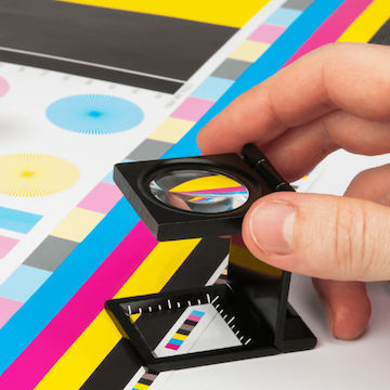 Hand holding small magnifying glass overtop some coloured bars printed on paper.